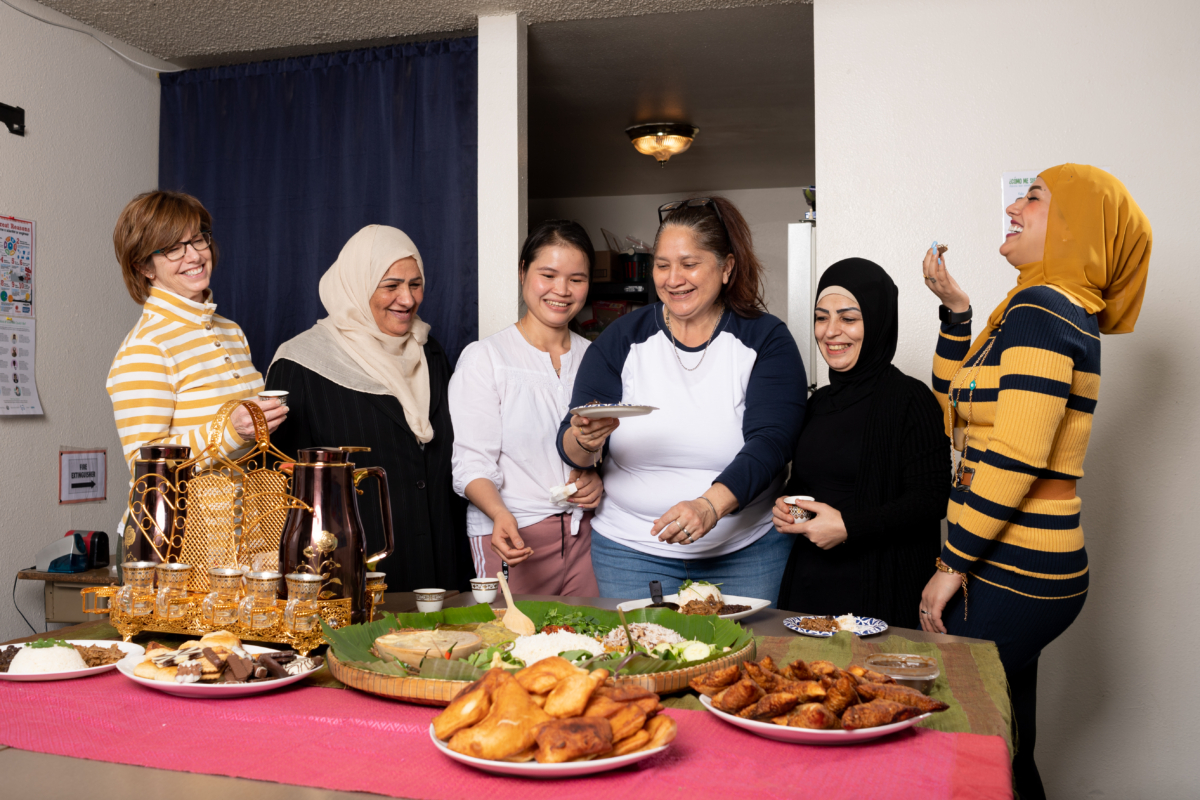 women from several cultures behind a table full of food. The women are laughing. Photo credit Kim Fetrow Photography